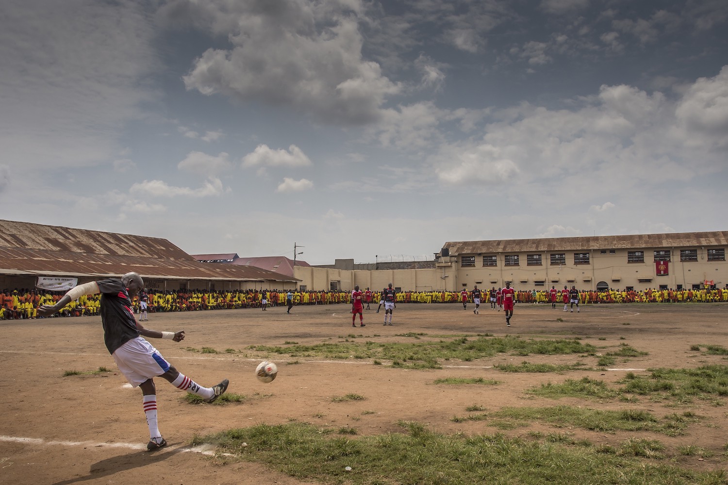 In Luzira Prison, in Kampala, a football league is organized. The teams composed of inmates meet each other throughout the year until a final. The teams are named mainly out of English Premier League clubs: Manchester United, Liverpool, Everton, ... The pitch is the courtyard of the Upper Prison, the most secure place of the Prison large compound. Hundreds of inmates come to support their favorite team. Liverpool in black versus Manchester United in Red. Liverpool will win the match.